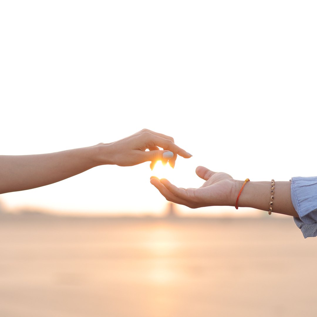 Two arms reaching toward each other against a bright sunset, fingertips almost touching, with the light glowing in the gap between them.
